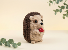 A crocheted hedgehog toy holding a small red strawberry, standing on a light beige surface against a soft cream background with decorative green leaves in the corners.