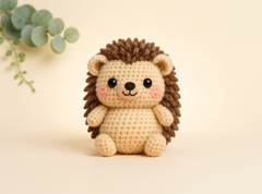 A crocheted hedgehog toy centered on a warm cream background, with a small green eucalyptus branch in the upper left corner. The hedgehog has a cream body, dark brown spines, black embroidered eyes and nose, and pink cheek accents.