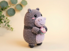 A crocheted hippo plush toy in dark gray and light lavender, holding a small pink flower, set against a warm beige background with soft green leaves in the corner.