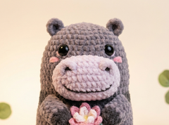 A crocheted hippo plush toy with a dark gray body, a pale pink snout and ears, black eyes, and a pink flower held in front, photographed against a soft cream background.