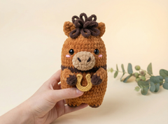 A hand holds a crocheted plush toy shaped like a moose or deer with a dark brown horn-like tuft on its head, a beige snout and inner ears, pink cheeks, and a yellow horseshoe accessory in its front hooves. The background is a soft cream color with a small eucalyptus sprig on the right.