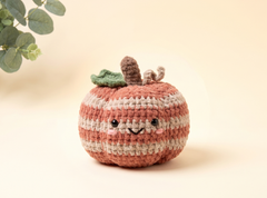 A cute crocheted pumpkin plush toy with a smiling face, featuring alternating terracotta and cream stripes, a green leaf, and a brown stem, set against a warm beige background with a soft leafy plant in the upper left corner.