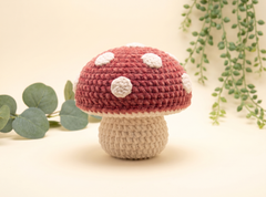 A crocheted mushroom decoration with a dusty rose red cap featuring white spots and a beige stem, styled with green eucalyptus and trailing plants on a warm cream background.