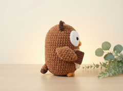 A crocheted owl plush toy in warm brown, sitting on a light wooden surface with a soft beige background and a sprig of green leaves to the right.