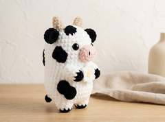 A crocheted cow plush toy with black and white spots, beige horns, and a pink snout, standing on a light wooden surface against a soft beige background with a blurred vase.