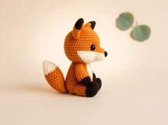 A crocheted fox toy sitting on a warm beige background with two green leaves in the upper right corner. The toy is primarily orange with white and black details.
