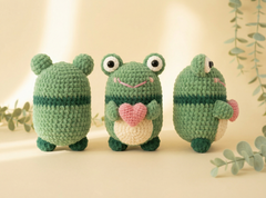 Three crocheted green frog toys arranged on a warm cream surface with soft leafy props. The center frog holds a pink heart, and all three have large white eyes and stitched facial details.