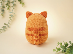 A crocheted orange cat-shaped amigurumi toy shown from the back, featuring pointed ears and a small tail, set against a warm cream background with soft green foliage accents in the corners.