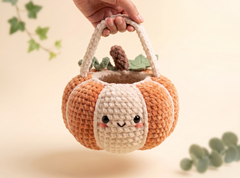 A crocheted pumpkin-shaped handbag with a cute face, held by a hand against a warm beige background with soft green leaf accents.