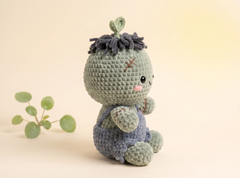 A crocheted plush doll with a round light sage-green head, dark charcoal yarn hair, and blue-gray overalls, posed on a warm beige background with a small green plant prop.