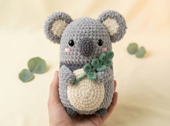 A crocheted koala plush toy held in a person's hand, featuring gray and cream yarn with green leaf accents, set against a soft beige background with decorative eucalyptus leaves.