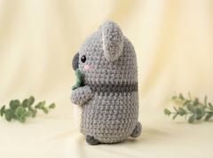 A crocheted koala plush toy in shades of gray and white, holding a green leaf, set against a soft beige background with decorative green leaves on the sides.