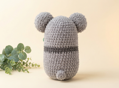 A gray crocheted plush toy shaped like a rounded character with two round ears on top and a small tail at the bottom, photographed on a warm cream background with a small green plant prop at the left edge.