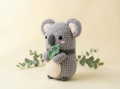 A crocheted koala toy holding a green leaf, standing against a warm beige background with decorative eucalyptus branches.