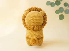 A crocheted plush lion toy shown from the back, made from textured mustard yellow yarn with a matching mane and pom-pom tail, set against a warm cream background with soft green eucalyptus leaves in the upper right.