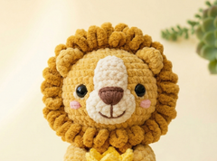 A close-up of a crocheted lion toy with a golden-brown mane, cream-colored face, pink cheeks, and a brown nose, set against a soft beige background with a blurred green plant in the corner.