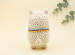 A crocheted plush toy shaped like a small bear or pig, shown from the back on a warm cream background with a decorative green leafy sprig in the upper right. The toy is cream-colored with a colorful striped band around its middle in red, yellow, and blue.