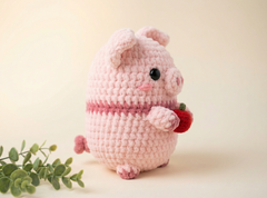 A pink crocheted plush pig toy holding a small red strawberry, photographed on a soft cream background with a sprig of green leaves at the lower left.