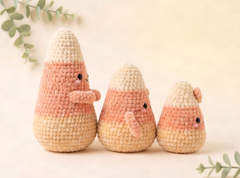 Three crocheted plush candy corn toys in different sizes, arranged on a soft cream background with subtle green leaf accents in the corners.