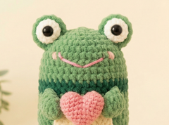 A green crocheted frog plush toy holding a pink heart, photographed against a soft cream background.