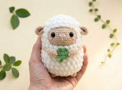 A small crocheted lamb toy held in a person's hand against a warm cream background with decorative green leaves. The lamb has a white textured body, tan face and ears, black bead eyes, pink cheeks, and a green four-leaf clover held in its front paws.
