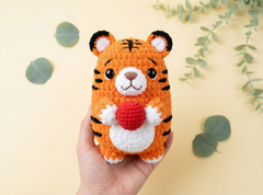 A hand holding a crocheted tiger plush toy with black stripes, a white face and belly, and a red heart-shaped accessory, set against a pale yellow background with decorative green leaves.