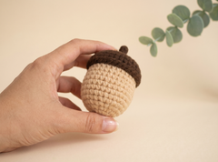 A hand holding a crocheted acorn ornament with a beige body and a dark brown cap, set against a warm beige background with a green leafy sprig in the upper right.