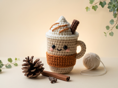Small amigurumi latte cup in beige and orange with black safety eyes and blush cheeks, on a cream backdrop.