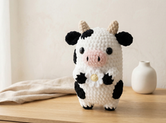 A crocheted cow figurine standing on a wooden surface, with a small white vase and draped fabric in the background.