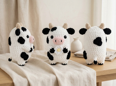 Three crocheted cow plush toys in black and white with beige horns and pink snouts, displayed on a wooden table draped with a cream linen cloth. One toy is shown from the front, and two are shown from the back, with a small white vase in the background.