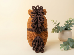 A crocheted horse-shaped item, likely a pouch or bag, shown from the back. It features a warm brown body with dark brown mane and tail details, set against a light beige background with a small potted eucalyptus plant on the right.
