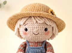 A crocheted doll with a beige straw hat, cream yarn hair, pink cheeks, and a blue overalls outfit over a patterned shirt, shown against a soft cream background.