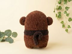 A brown crocheted plush toy shaped like a small bear or animal, shown from the back on a warm cream background with decorative green leaves and ivy.