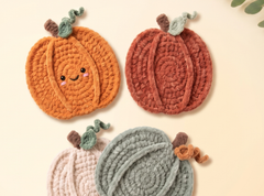 Three crocheted pumpkin-shaped items arranged on a light cream background: an orange one with a smiling face and green stem, a rust-orange one, and a pale pink one partially visible at the bottom along with a muted blue-gray one also partially visible. A small leaf appears in the top-right corner.