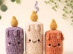 Three cute crocheted candle characters in purple, cream, and orange, each with embroidered faces and small yarn flames on top, set against a soft beige background with blurred green leaves.