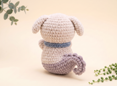 A crocheted plush dog toy shown from the back, featuring a cream-colored body, floppy ears, a blue collar, and a lavender tail, set against a warm beige background with decorative green plant sprigs.