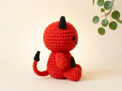 A red crocheted devil doll with black horns, tail tips, and ears, sitting on a light beige background with a green plant leaf in the upper right corner.