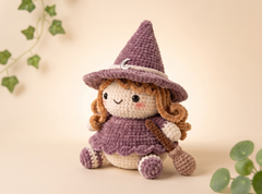 A crocheted witch doll with a purple hat and dress, cream face and belly, and brown curly hair, posed on a warm beige background with soft green ivy leaves in the corners.