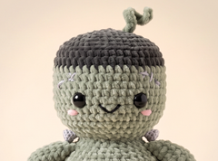 A close-up of a crocheted plush doll with a light sage green body, a dark charcoal gray top, and small pink embroidered cheeks, set against a solid beige background.