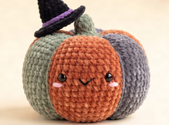 A cute hand-crocheted pumpkin doll with a black witch hat, featuring orange, gray, and sage green yarn sections, embroidered facial features, and a small stem, set against a warm beige background.