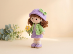 A crocheted doll with curly brown hair, wearing a lavender hat with a green bow, a green cardigan, and a lavender dress, holding a yellow flower. The doll is posed on a warm beige surface with soft-focus green leaves in the background.