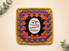 A square crocheted granny square featuring a mummy face motif, surrounded by concentric rings of orange, purple, olive green, and black yarn, finished with a mustard yellow border. The square is displayed flat on a light beige background with decorative eucalyptus leaves in the corners.