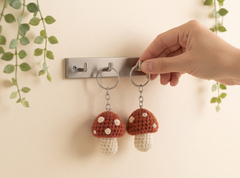 A hand holds a silver keyring with a crocheted mushroom-shaped keychain against a beige wall with a metal hook and green trailing leaves.