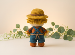 A crocheted scarecrow doll shown from the back, wearing a yellow hat with a brown band, a white face, orange and blue overalls, and brown shoes. It is styled with green eucalyptus branches on a warm beige background.