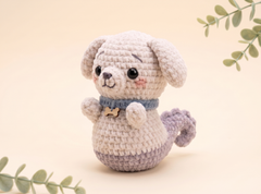 A crocheted plush dog toy with a cream-colored body, lavender belly and tail, and a blue collar with a small bone-shaped charm, set against a warm beige background with decorative green leaves.