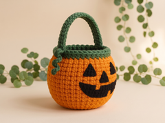 A crocheted pumpkin-shaped candy bucket with a green handle and trim, featuring a black stitched jack-o'-lantern face, set against a warm beige background with decorative green eucalyptus leaves.
