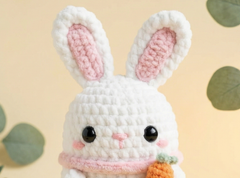 A close-up of a crocheted white bunny plush toy with pink inner ears, black bead eyes, a pink nose and scarf, and a small orange carrot, set against a soft beige background with blurred green leaves.