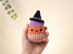 A hand holds a cute, crocheted plush toy shaped like a candy corn wearing a witch hat. The toy has a purple top section, an orange middle section, and a pale pink bottom section, with a black pointed hat.