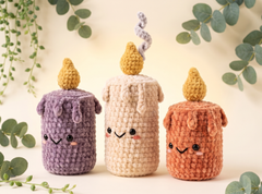 Three cute crocheted candle-shaped plush toys with embroidered smiling faces are arranged on a light cream surface with green eucalyptus leaves around them. The candles are in purple, cream, and terracotta orange, each with a yellow flame on top.
