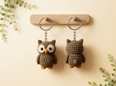Two crocheted owl keychains in brown are displayed on a wooden rack against a warm beige background, with decorative green leaves in the corners. One keychain is shown from the front and the other from the back.